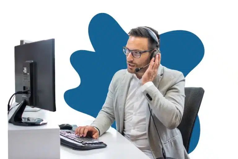 A male call center agent wearing a gray blazer and glasses, speaking into a headset while working on a desktop computer. The background features a blue abstract shape.