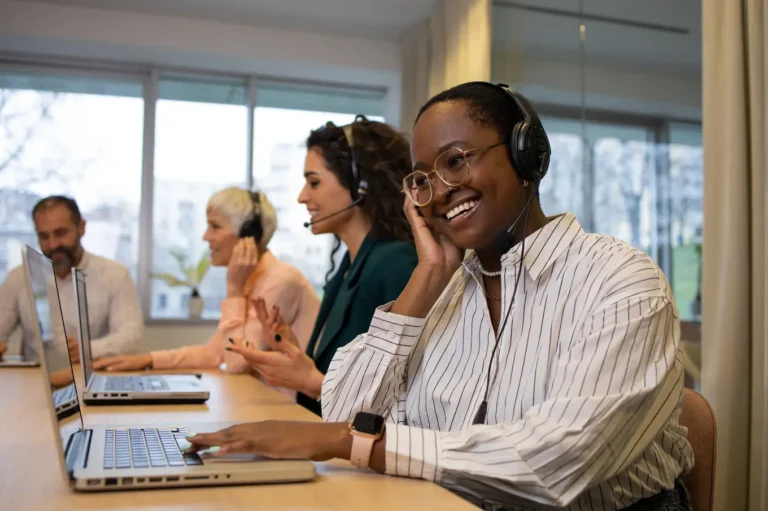 A diverse group of call center agents sitting at a table, smiling and wearing headsets while working on laptops. A bright and modern office space is visible in the background.