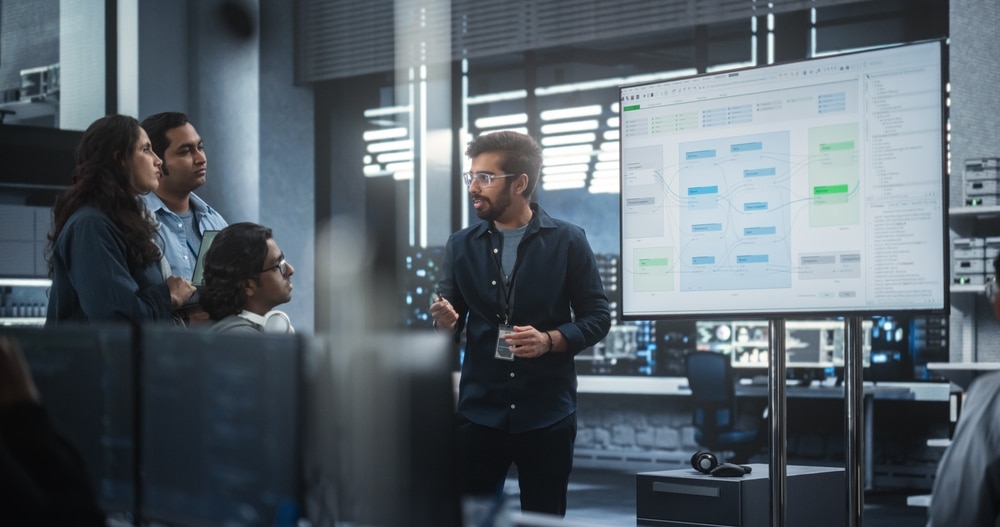 A professional presenting a data workflow diagram on a digital whiteboard to a group of colleagues in a modern office setting, symbolizing team collaboration and technological planning.