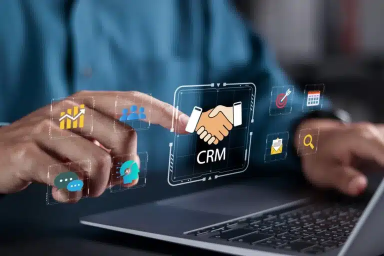 A person interacts with a virtual screen displaying CRM icons, including handshake, chat, and graphs, over a laptop, indicating customer relationship management.