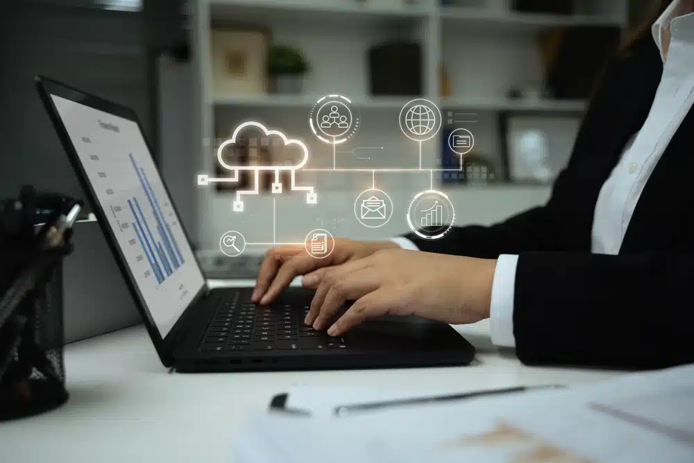A person in a suit types on a laptop with cloud technology icons hovering above. A chart on the screen indicates data analysis. Office setting, professional tone.