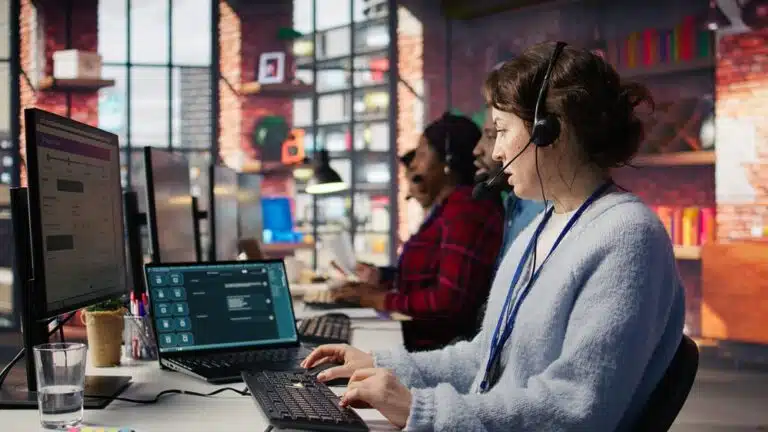 Three focused call center employees work at computers with headsets in a bright, modern office. Large windows and colorful decor create an energetic atmosphere.