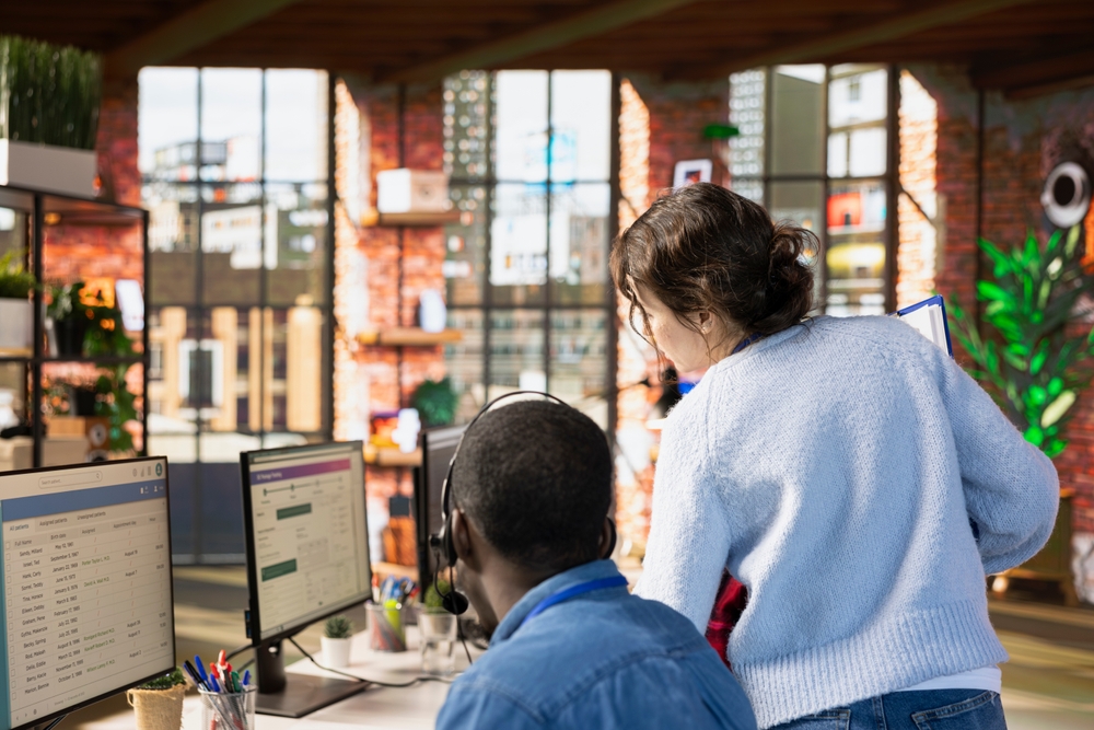 Two people collaborating on computers in an office, discussing Xima vs Custom-Built Contact Center Solutions for small businesses.