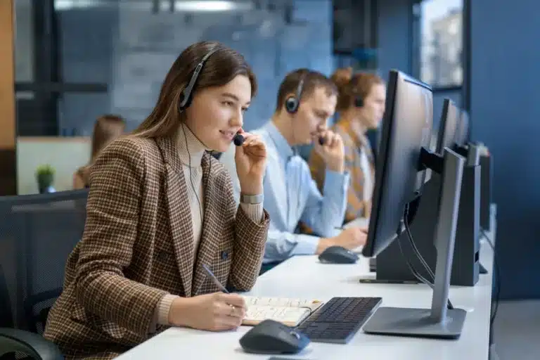 Customer service representatives wearing headsets working at desktop computers in a modern call center office, with one agent taking notes while assisting customers.