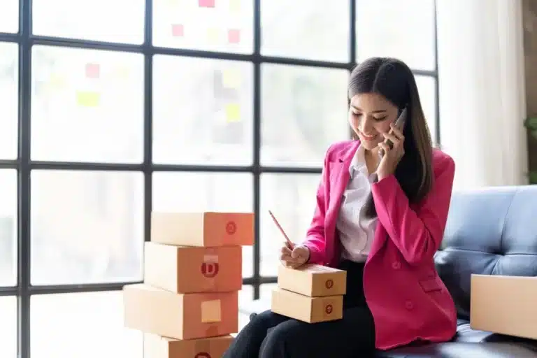 A woman in a pink suit sits on a couch, talking on the phone surrounded by unpacked boxes.